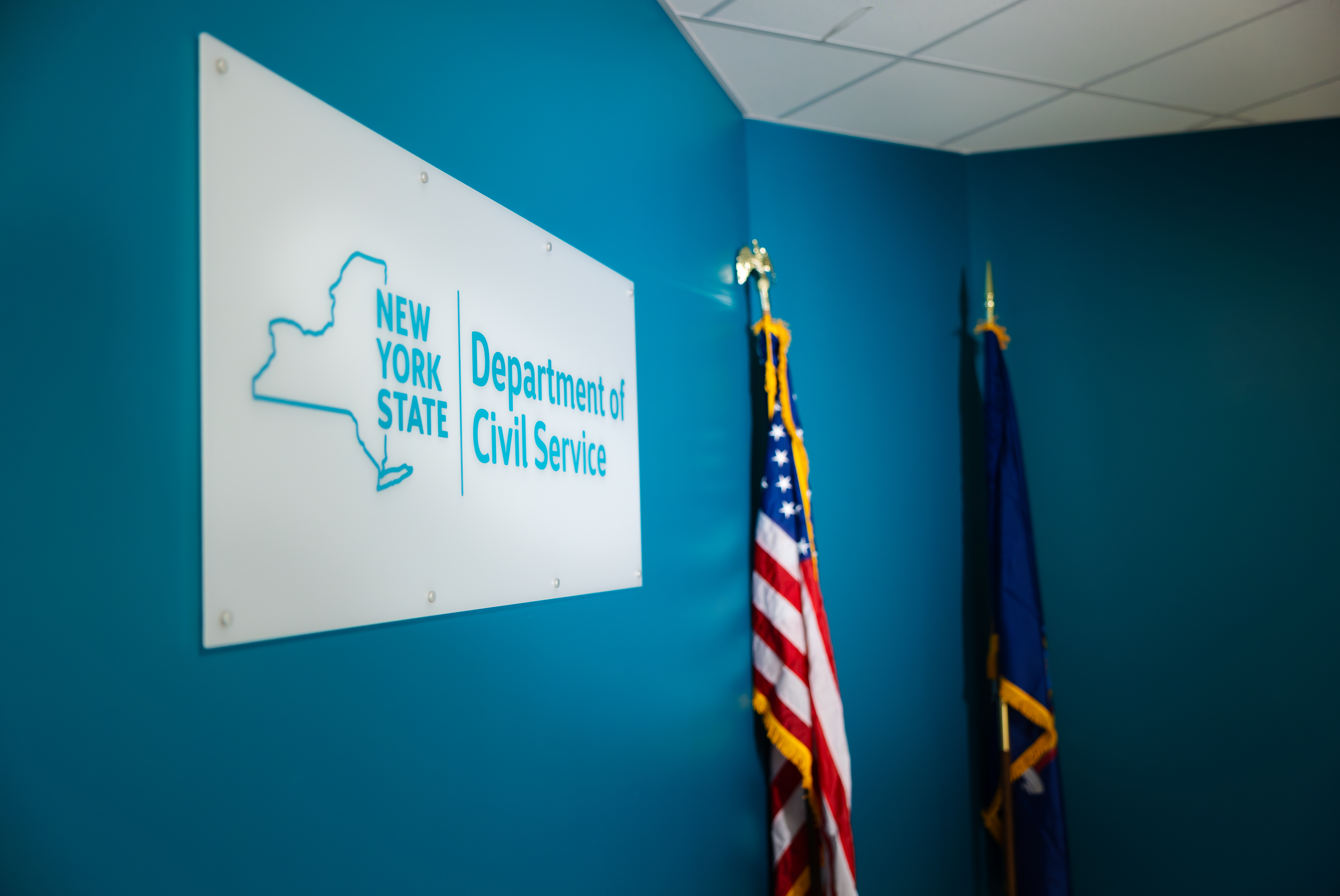 New York State Department of Civil Service sign mounted on the wall with American and New York State flag in the back.